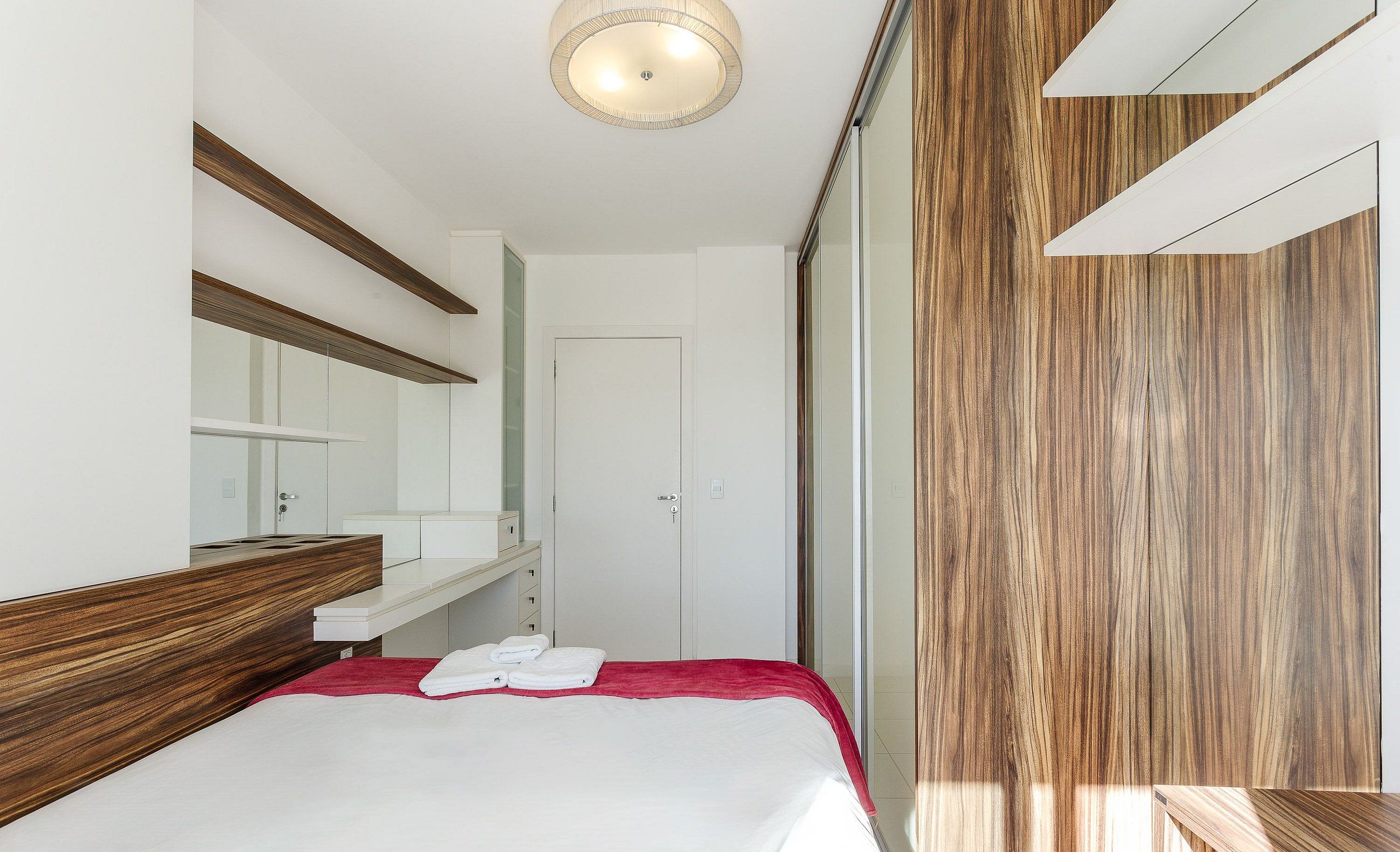 A modern hotel room with a wooden headboard, a large mirror, a white bed with a red accent, and a sliding door leading to a shower area.