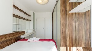 Una habitación de hotel moderna con un cabecero de madera, un espejo grande, una cama blanca con un detalle rojo y una puerta corredera que da a una zona de ducha.