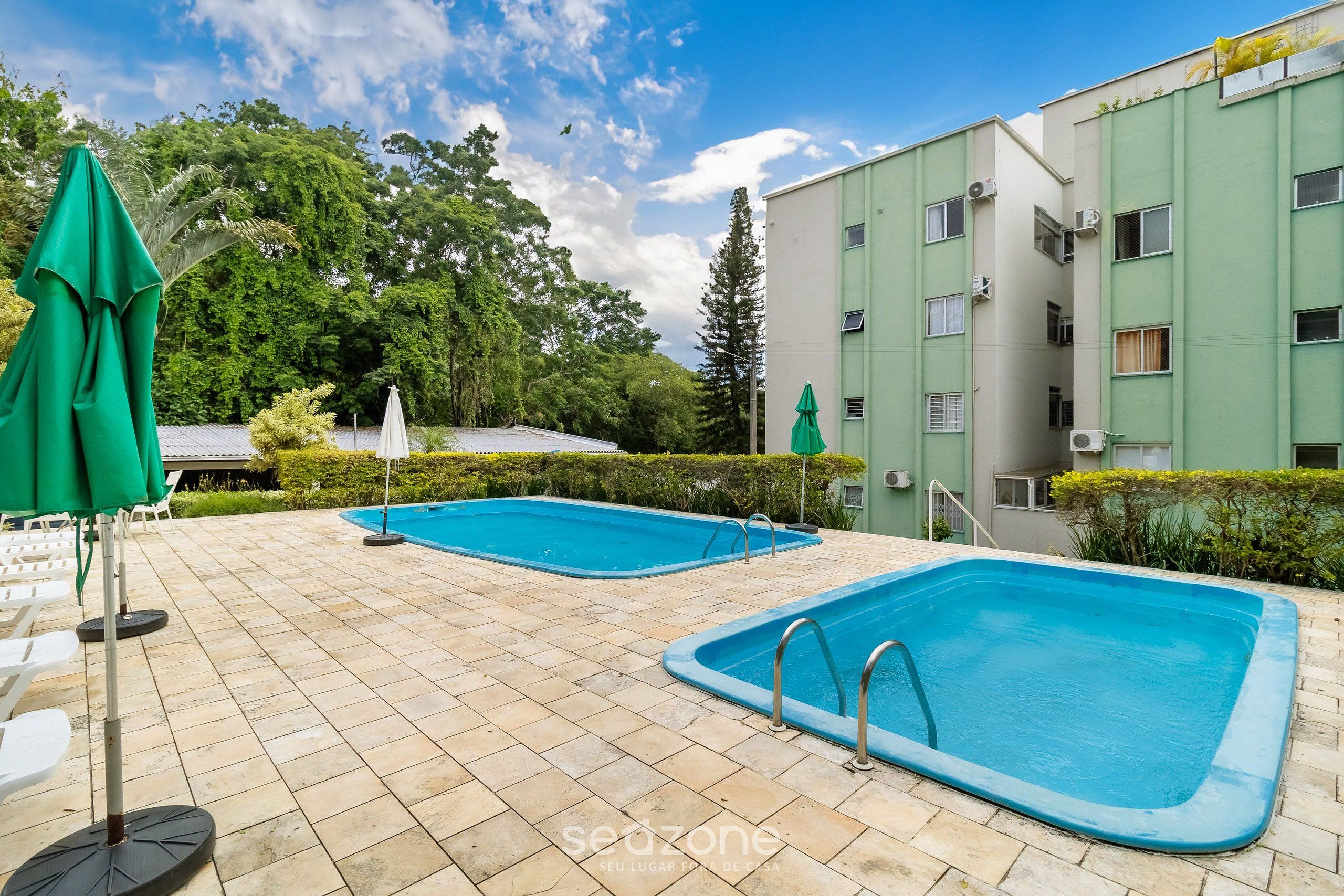 Un área de piscina con dos piscinas, rodeada por un solárium con baldosas, y un edificio con múltiples ventanas y unidades de aire acondicionado.