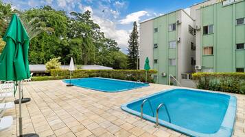 Un área de piscina con dos piscinas, rodeada por un solárium con baldosas, y un edificio con múltiples ventanas y unidades de aire acondicionado.