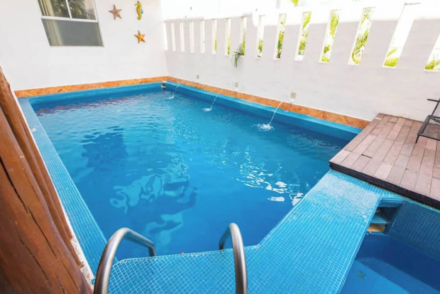 Une piscine dont le bord est recouvert de carreaux bleus, avec une terrasse en bois et un mur blanc orné de motifs en forme d’étoiles.
