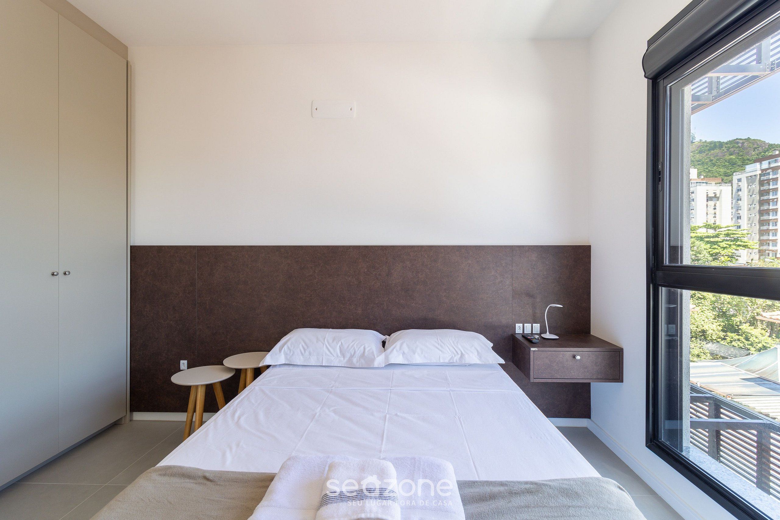 Un dormitorio moderno con una cama grande, mesitas de noche, un ventanal con vistas a edificios y un armario.