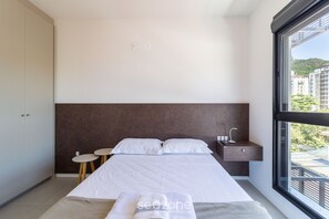Un dormitorio moderno con una cama grande, mesitas de noche, un ventanal con vistas a edificios y un armario.