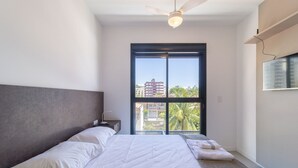 A bedroom with a large window, a ceiling fan, and a bed with white linens. - Modern Studio 650m From Ufsc - Cnt303 (Florianópolis)