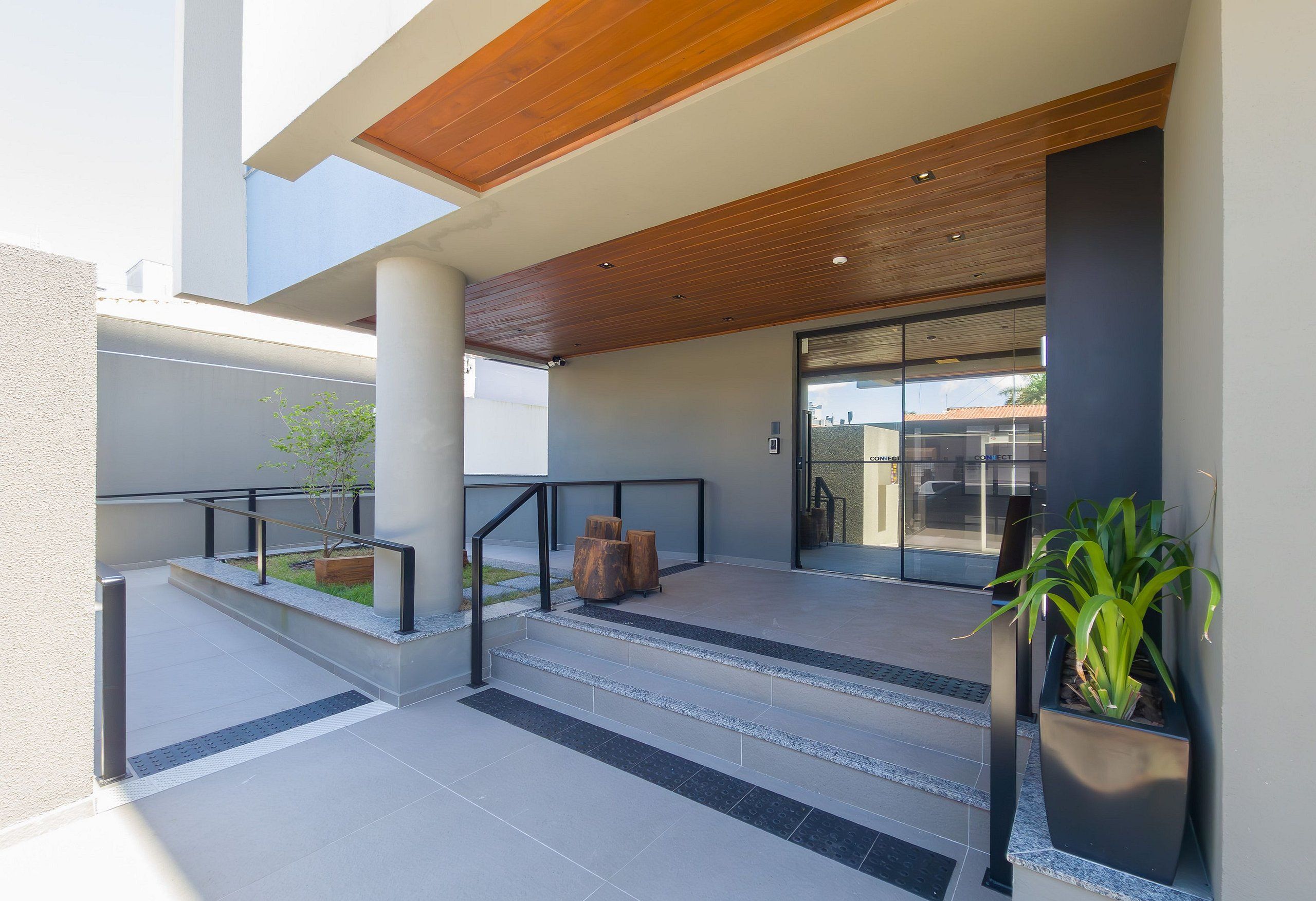 Entrada de un edificio moderno con techo de madera, puerta de cristal y un pequeño área de descanso al aire libre con escalones y una jardinera.