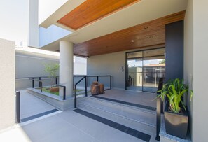 Entrada de un edificio moderno con techo de madera, puerta de cristal y un pequeño área de descanso al aire libre con escalones y una jardinera.