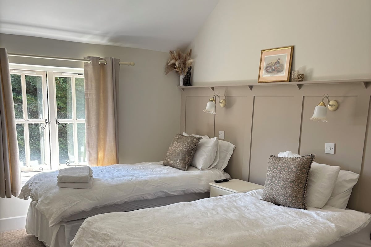 A bedroom with two beds, a window with curtains, a wall-mounted lamp, and a framed picture.