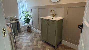 A bathroom with a herringbone-patterned wooden floor, a green cabinet with a gold faucet, and a round mirror on the wall.