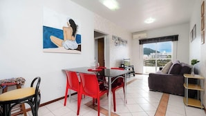 A modern living room with a dining area, featuring a large window with a view of buildings and a balcony.