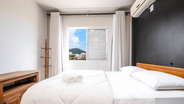 A hotel room with a bed, a wooden nightstand, a window with curtains, and a view of buildings and a hill.