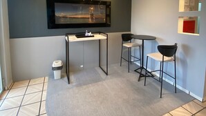 Een moderne kamer met een grote flatscreen televisie aan de muur, een kleine tafel met twee stoelen, een prullenbak en een betegelde vloer.