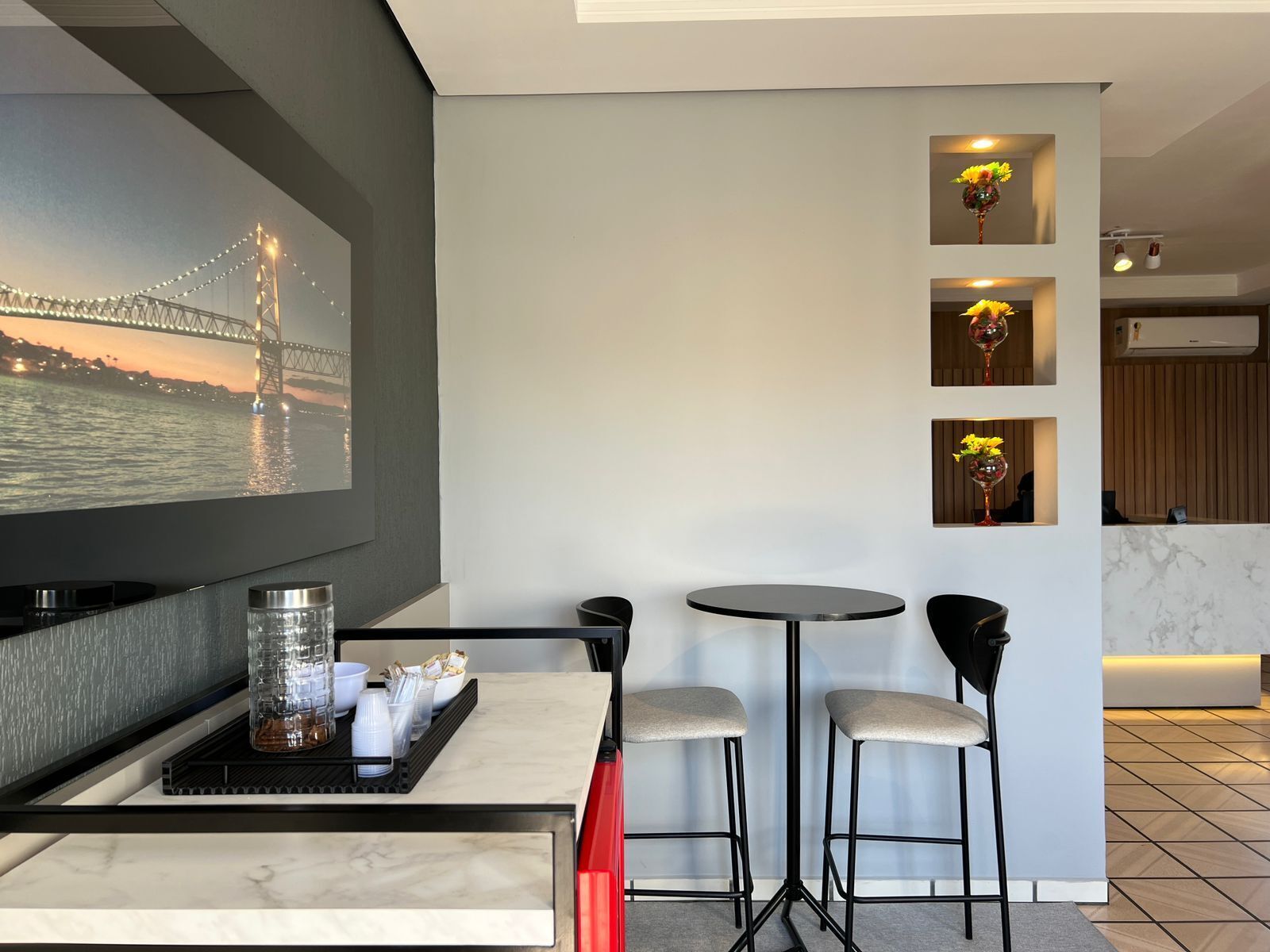Habitación moderna con un televisor grande que muestra un paisaje urbano, una pequeña zona de comedor con una mesa y dos taburetes altos, y una estantería de pared con adornos.