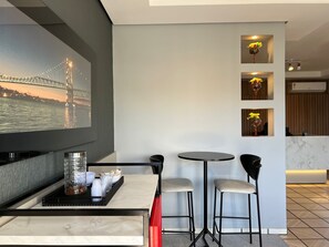 Habitación moderna con un televisor grande que muestra un paisaje urbano, una pequeña zona de comedor con una mesa y dos taburetes altos, y una estantería de pared con adornos.