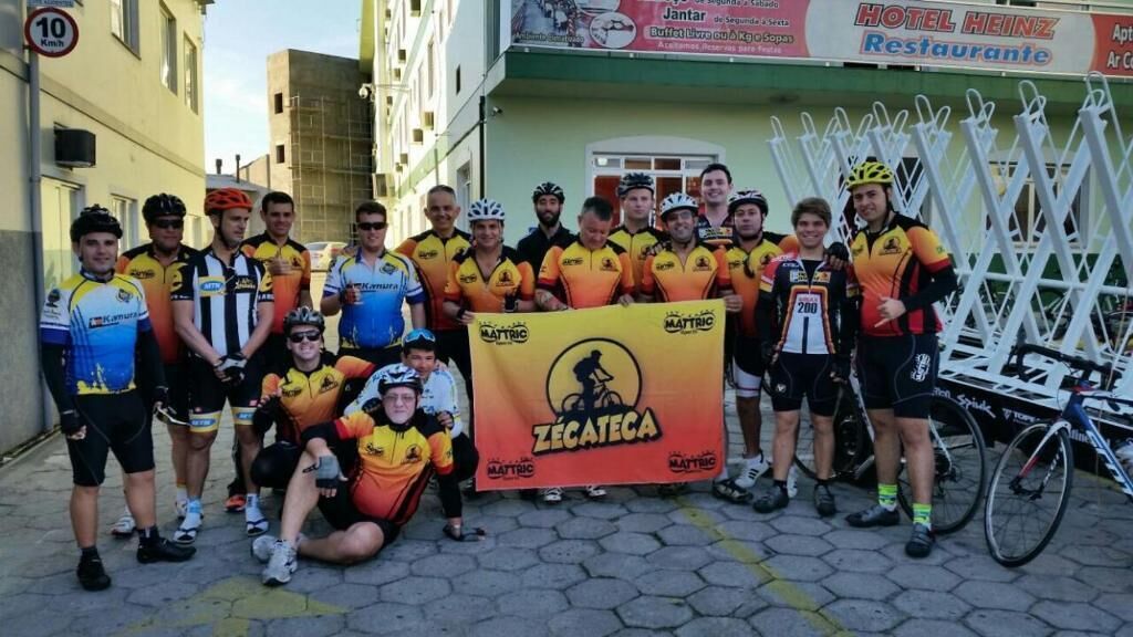 A group of cyclists posing with a banner in front of a building with a sign for Hotel Heinz Restaurante.