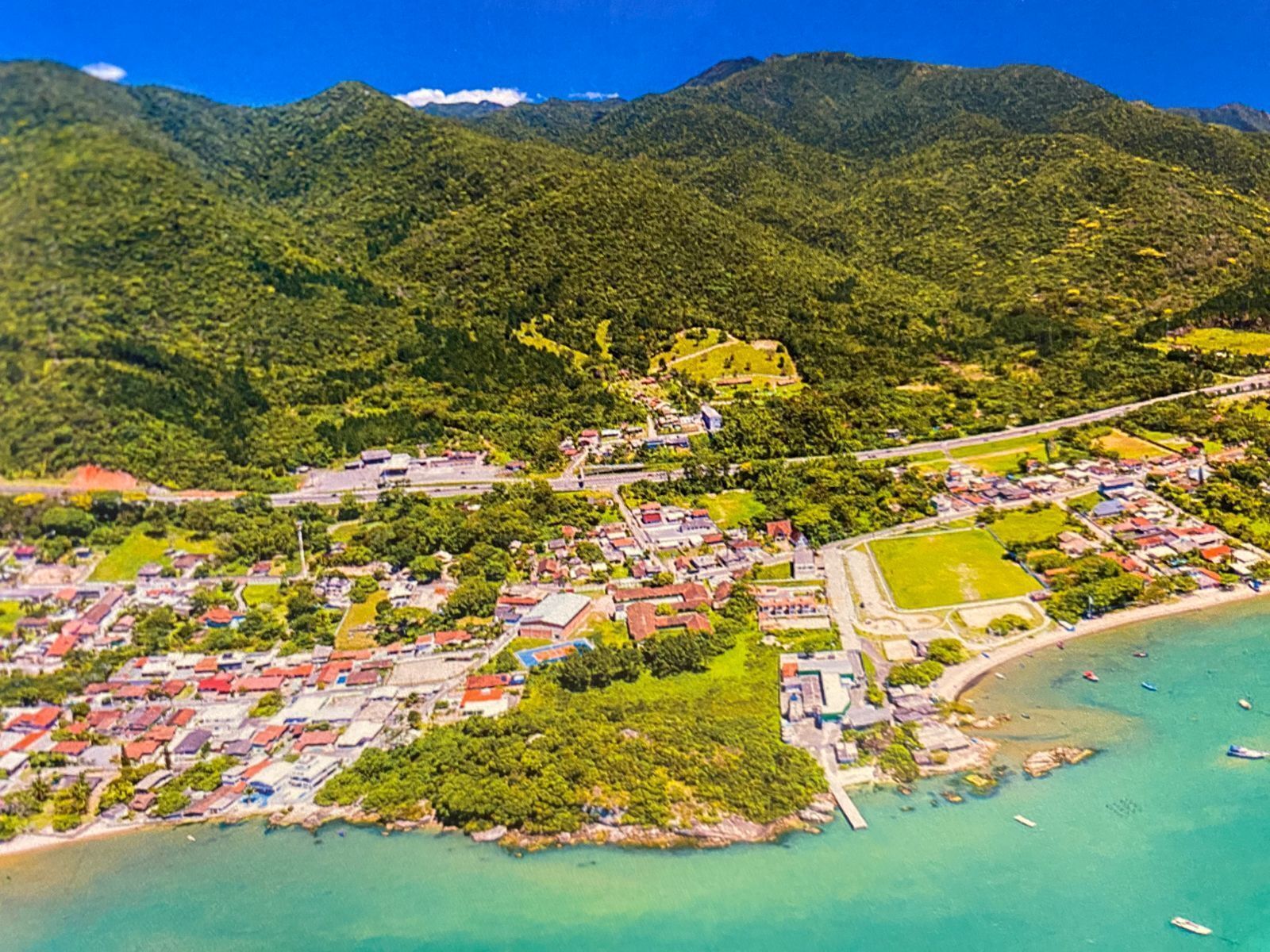 Une vue aérienne d’une ville côtière, avec un mélange de bâtiments résidentiels et commerciaux, une imposante structure ressemblant à un château et une plage où se trouvent des bateaux.