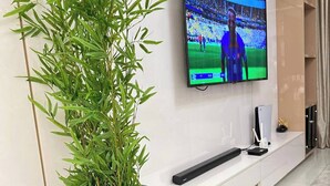 A modern living room with a flat-screen TV mounted on the wall, a black plant in a pot, and a minimalist entertainment unit with a remote control. - Cozy Apartments By Signature (Abuja)