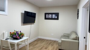 Een kamer met een flatscreen televisie aan de muur, een beige bank, een witte tafel met een bloemenvaas, en een houten vloer.