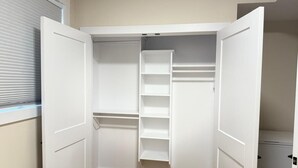 Een witte inloopkast met meerdere planken en knaakruimte, in een kamer met houten vloer en een raam met zonwering.