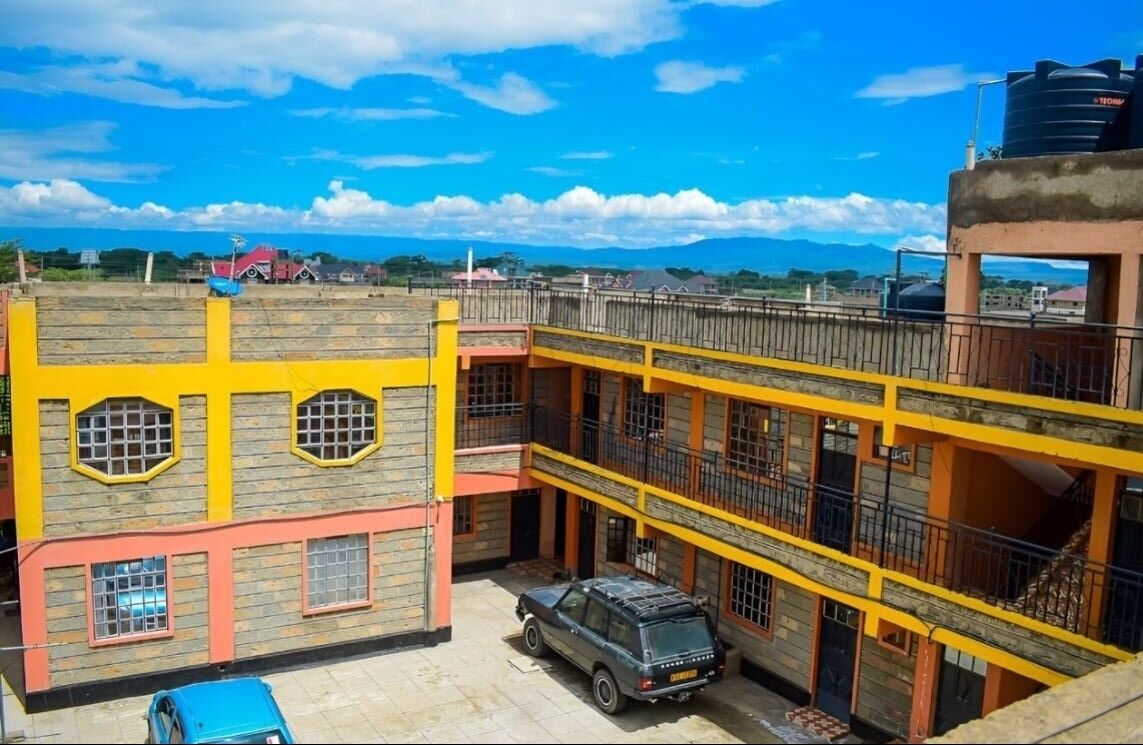 Un edificio de varios pisos con fachada amarilla y roja, un coche estacionado adelante y un cielo azul despejado.