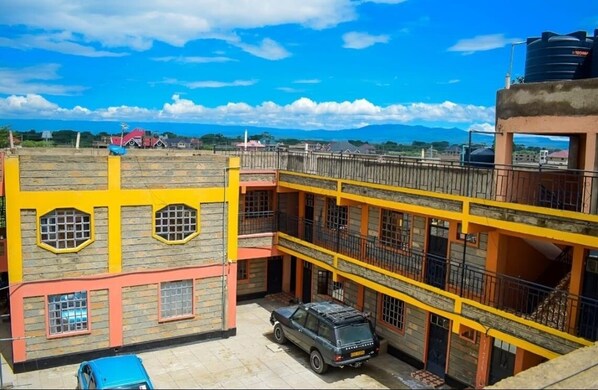 A multi-story building with a yellow and red facade, a car parked in front, and a clear blue sky. - Cozy Naivasha Apartment With Mountain Views (Naivasha)