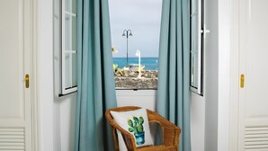Ein Zimmer mit Meerblick durch ein Fenster, ein Korbstuhl mit Kaktus-Kissen und eine Steinmauer draußen.