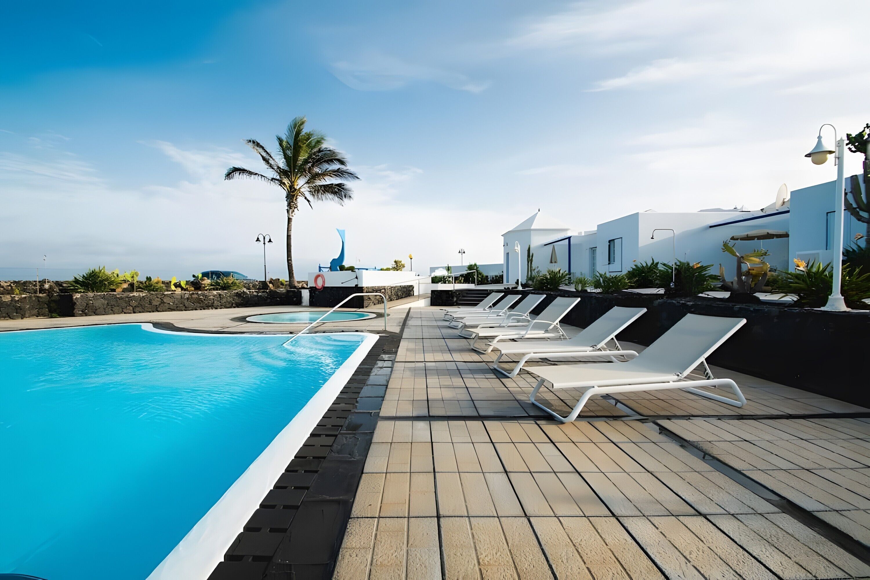 A pool area with lounge chairs, a palm tree, and a building with a white facade.