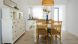 Un comedor con sillas de mimbre, un armario blanco con cajones, una lámpara colgante de mimbre y un jarrón con flores sobre la mesa.