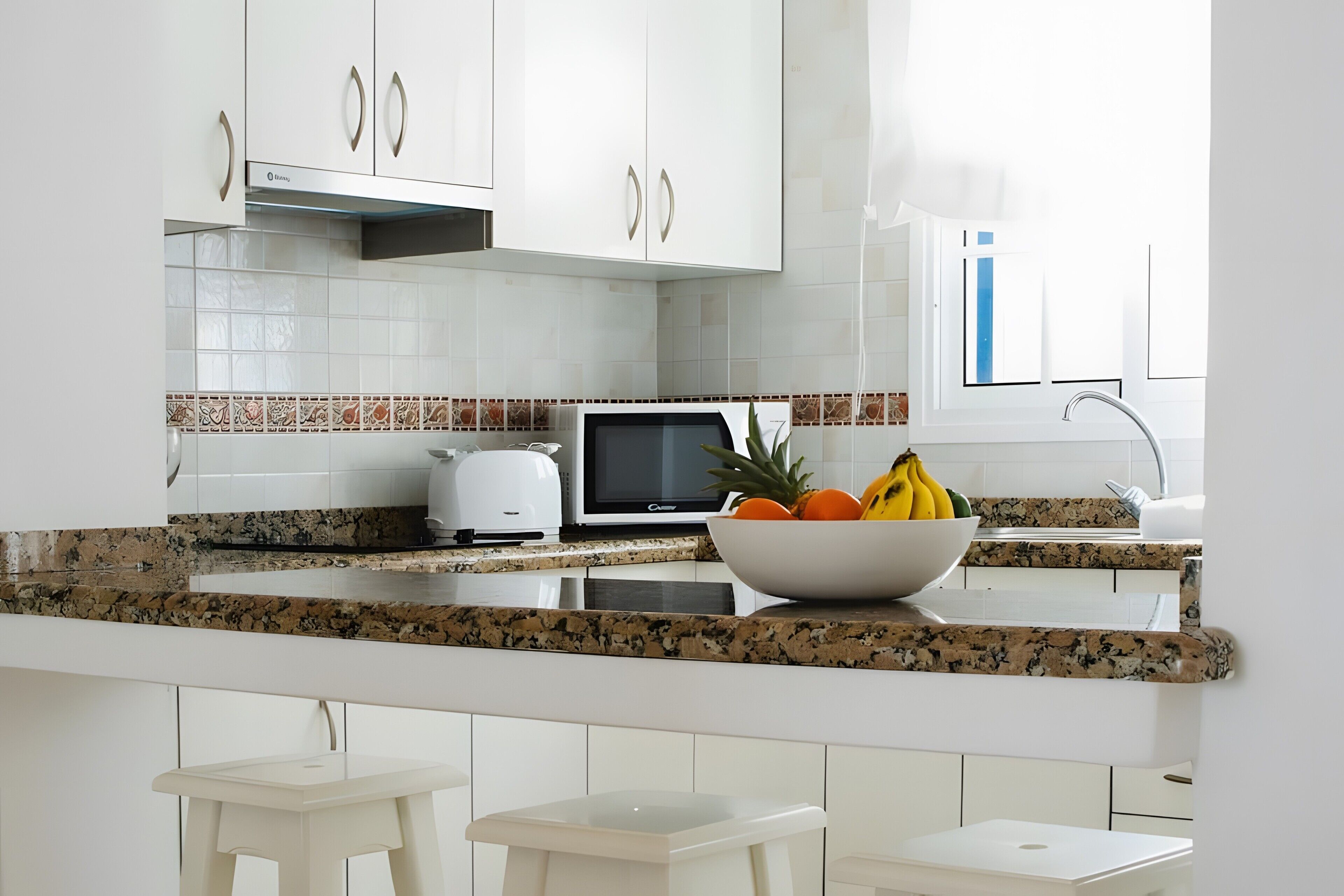 Una cucina moderna con mobili bianchi, un piano di lavoro in granito, un forno a microonde e una ciotola di frutta.