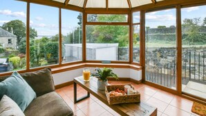 A conservatory with a cushioned bench, a table with a drink, a basket of bread, and a potted plant. - Blacksands Seaview Cottage, Aberdour (Aberdour)