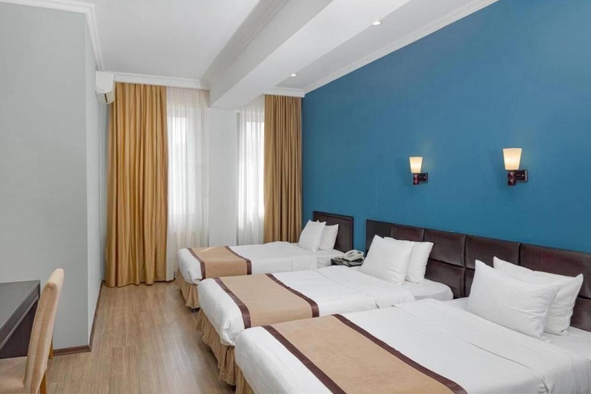 Hotellihuoneessa on kolme sänkyä, työpöytä ja tuoli. Huoneessa on kaksi seinään kiinnitettyä lamppua ja ikkuna, jossa on verhot.