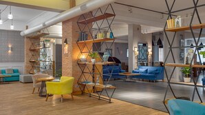 A spacious lobby with a brick column, wooden shelves, and a mix of seating areas including a yellow armchair and blue sofas. - Hotel Mevora (Tbilisi)