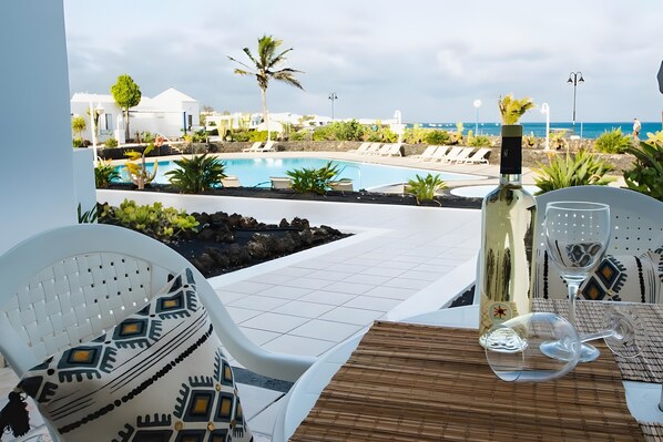 A poolside table with a bottle and glasses, overlooking a pool and lounge area with palm trees and a view of the sea. - Holiday Home 'Caleta Campos Bungalows' with Sea View, Shared Pool and Wi-Fi (Arrieta)