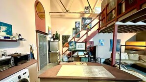 A compact kitchen with a washing machine, a microwave, and a small refrigerator. There is a wooden counter with a cutting board and a sink. The kitchen is adjacent to a living area with a couch and a staircase leading to an upper level. - Arty Infused Loft With ac Near Emirates Stadium (London)