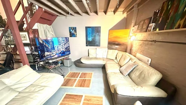 A modern living room with a sectional sofa, a flat-screen TV, and a wooden floor. - Arty Infused Loft With ac Near Emirates Stadium (London)