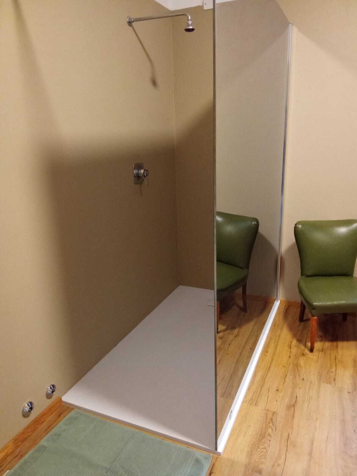 Una habitación con piso de madera, una silla verde y un área de ducha con puerta de vidrio.