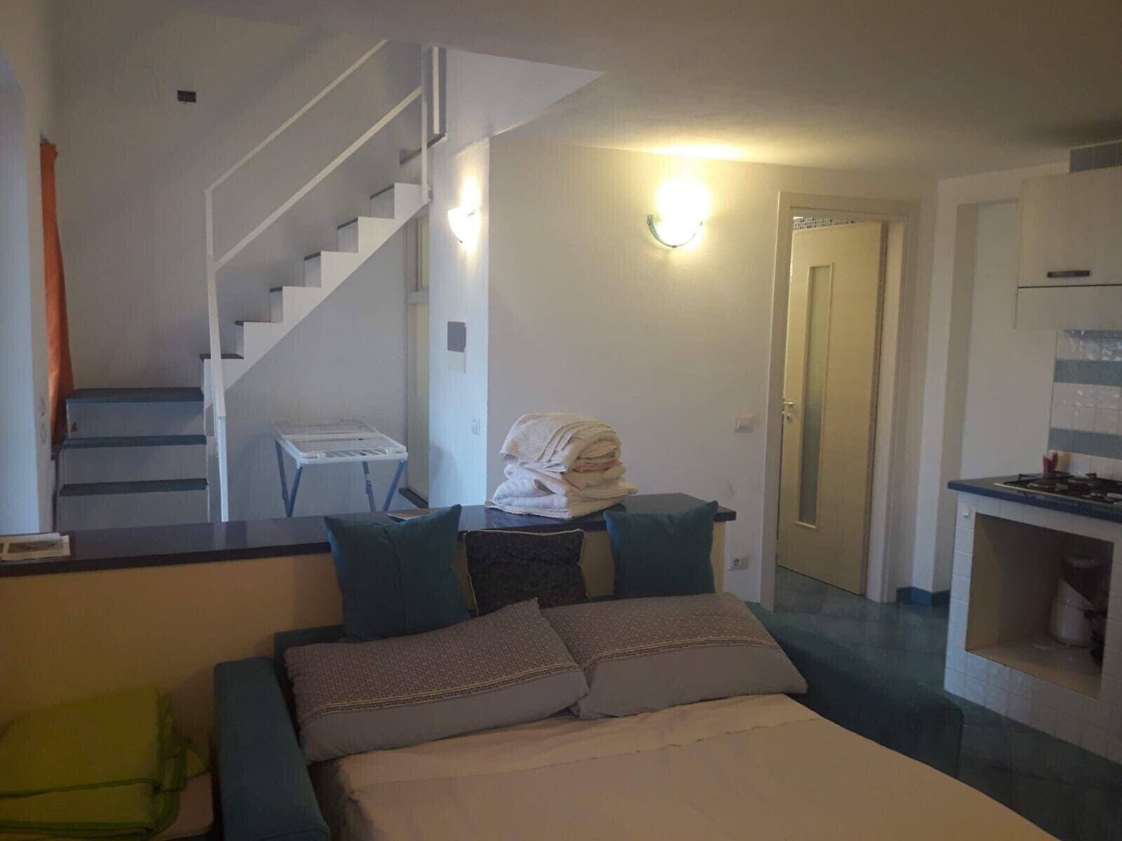 A bedroom with a bed, pillows, a desk, a chair, a staircase, and a kitchen area.