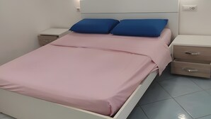 Ein Bett mit rosa Bettdecke und blauen Kissen, ein weißes Kopfteil, zwei Nachttische mit Schubladen und ein Lichtschalter an der Wand.