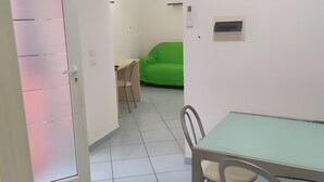 Ein Zimmer mit Esstisch, Stühlen, einem grünen Sofa, einer weißen Tür und einem gefliesten Boden.