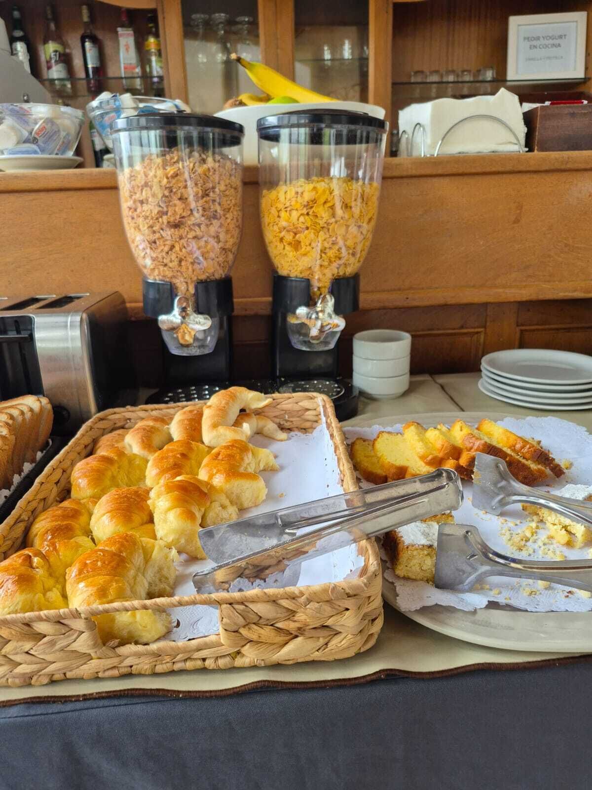 A buffet setup with bread rolls and a bread basket, along with two large dispensers of cereal.
