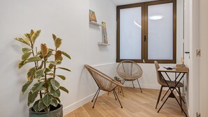 A room with a large window, a potted plant, two wicker chairs, a small table, and a wall-mounted shelf with pictures.