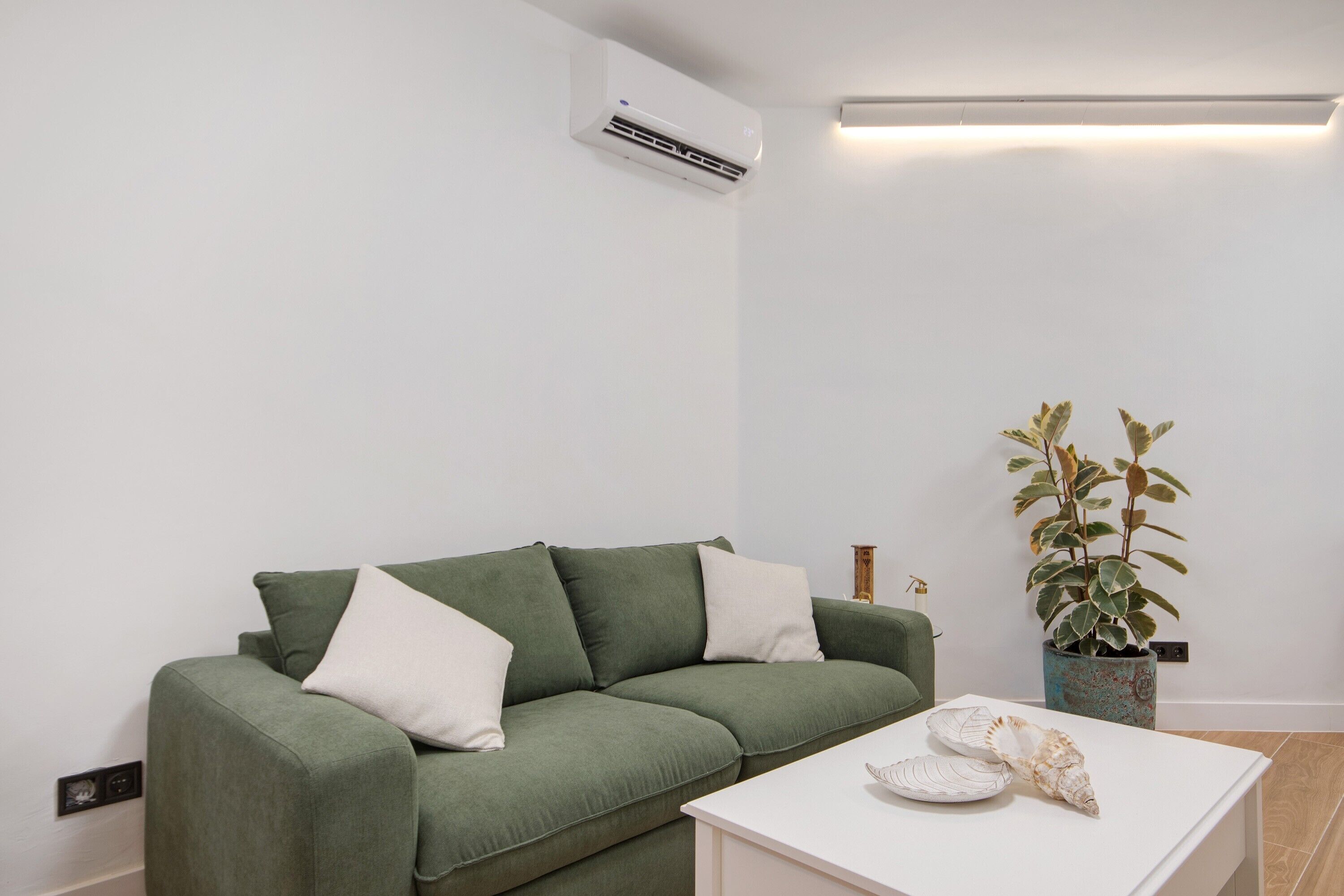 A green sofa with white cushions, a white side table with decorative shells, a potted plant, and a wall-mounted air conditioner.