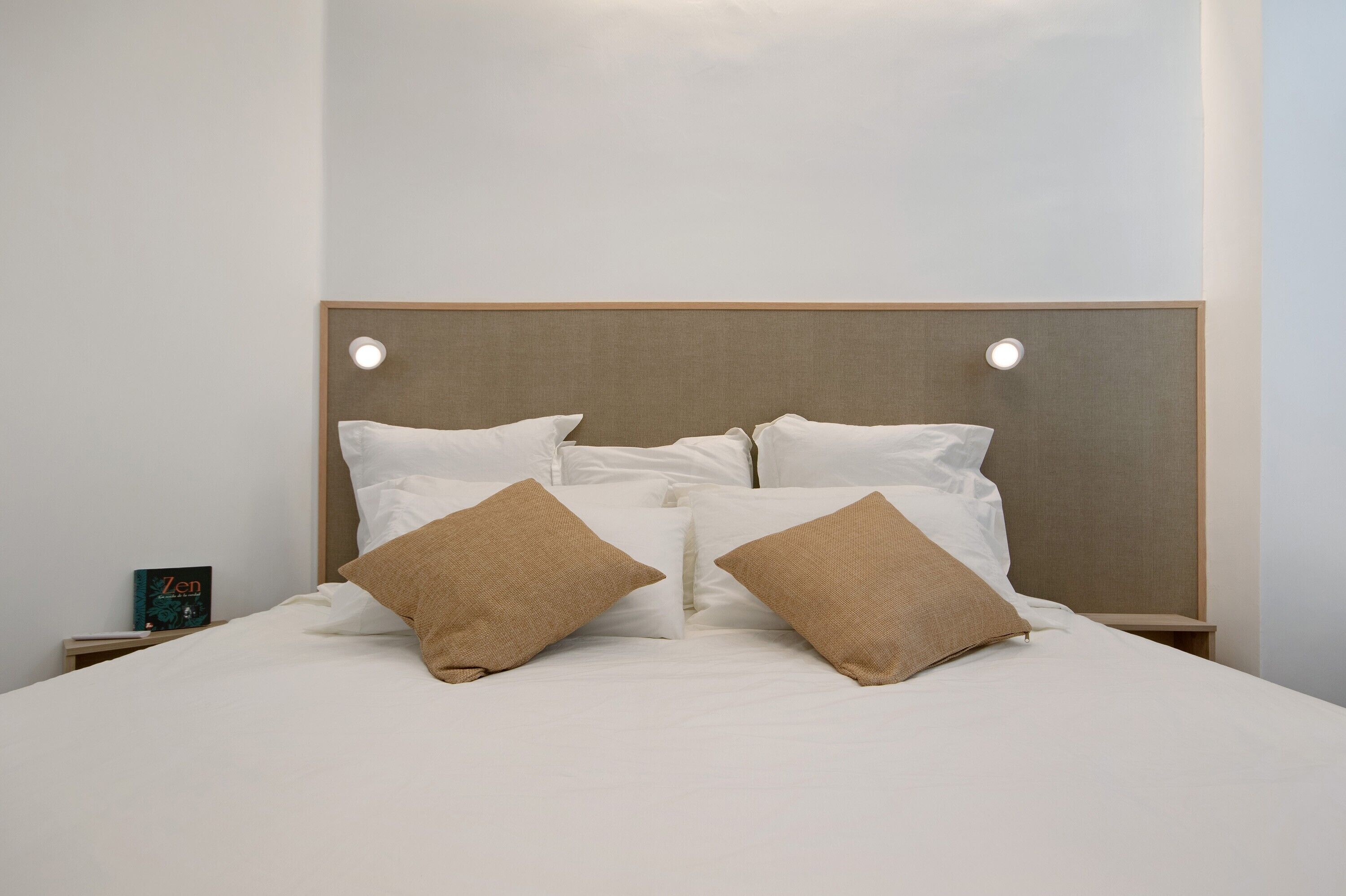 Un letto rifatto con lenzuola bianche e due cuscini marroni, un comodino con un libro e un lampadario fissato alla parete.