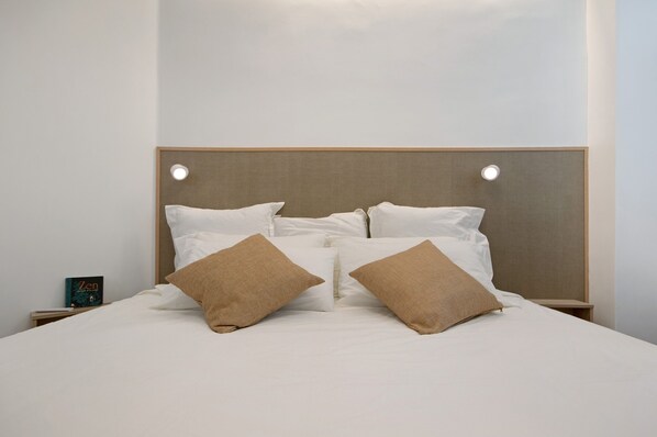 A neatly made bed with white bedding and two brown pillows, a bedside table with a book, and a wall-mounted light fixture.