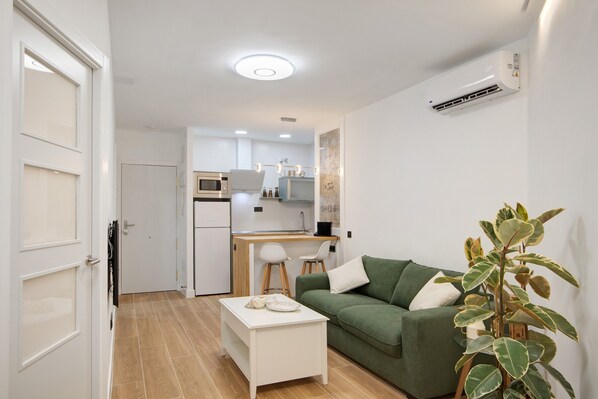 A compact living space with a kitchenette, a green sofa, a white coffee table, and a potted plant.