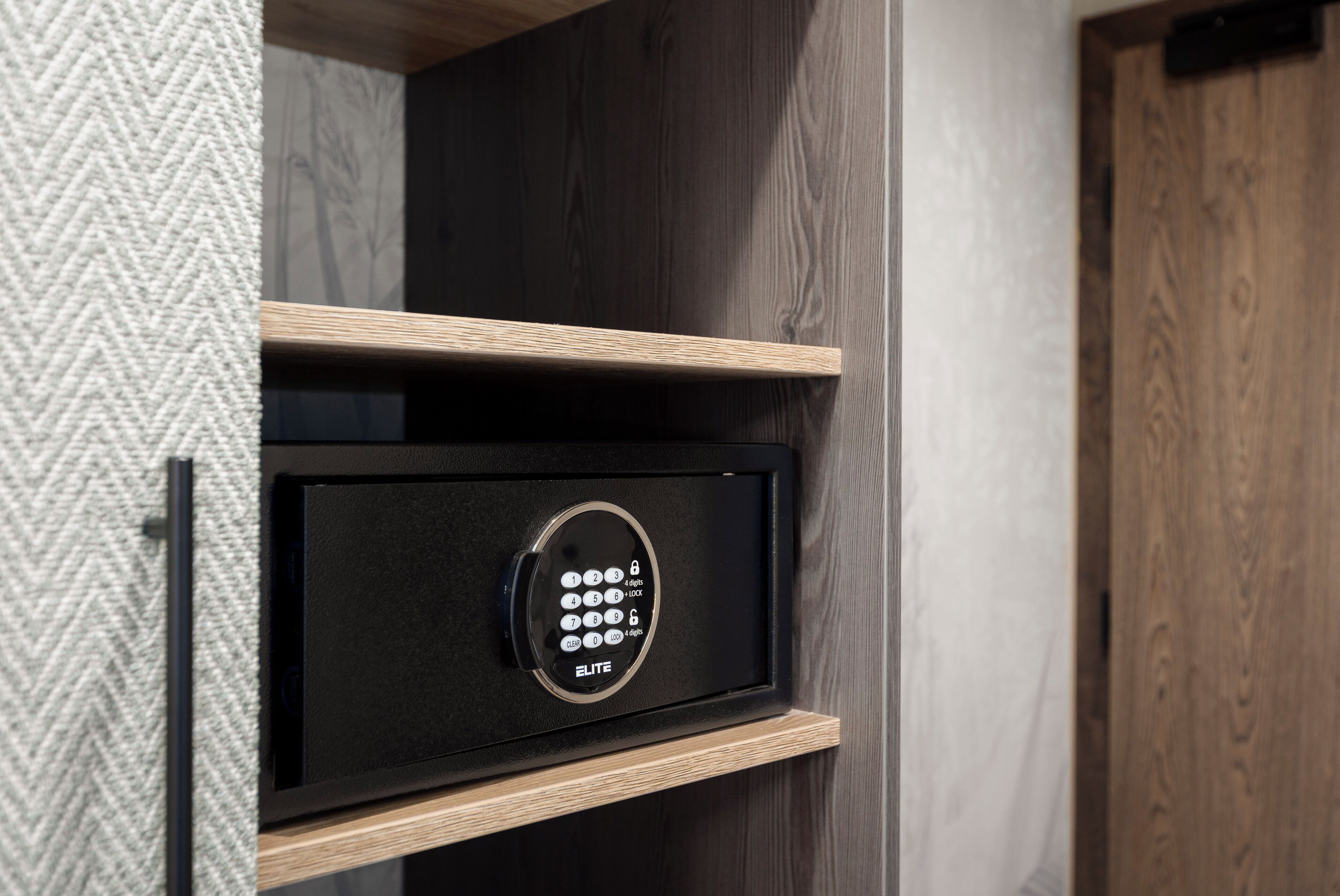 A black safe with a keypad, labeled "ELITE," mounted on a wooden shelf in a room with a herringbone-patterned wall.