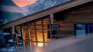 Un chalet en bois doté de grandes fenêtres, d’un balcon et offrant une vue sur les montagnes enneigées au coucher du soleil.