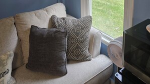 Una habitación con pared azul, una ventana con persianas, un sofá con cojines estampados, un cuadro y un televisor.
