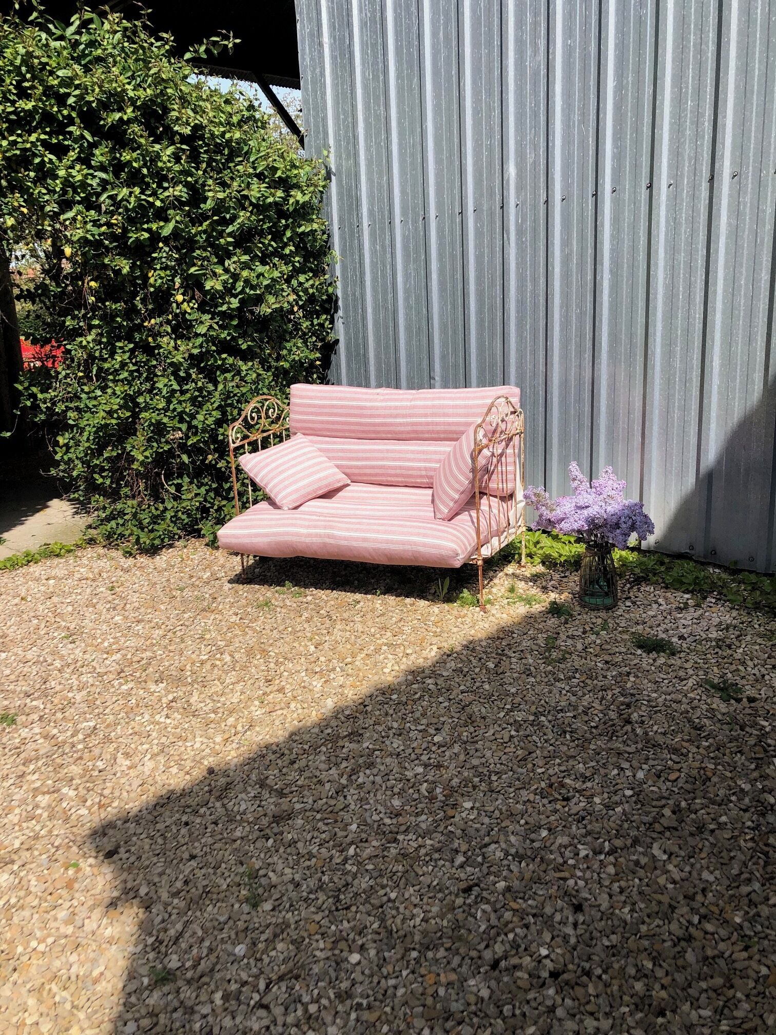 A pink striped sofa with a cushion, positioned on a gravel surface, with a metal fence and a bush in the background.
