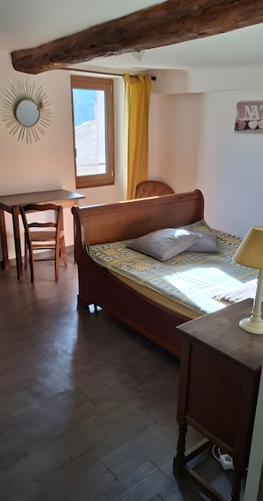 Quarto com cama de madeira, escrivaninha, cadeira, janela com cortinas e espelho fixado na parede.