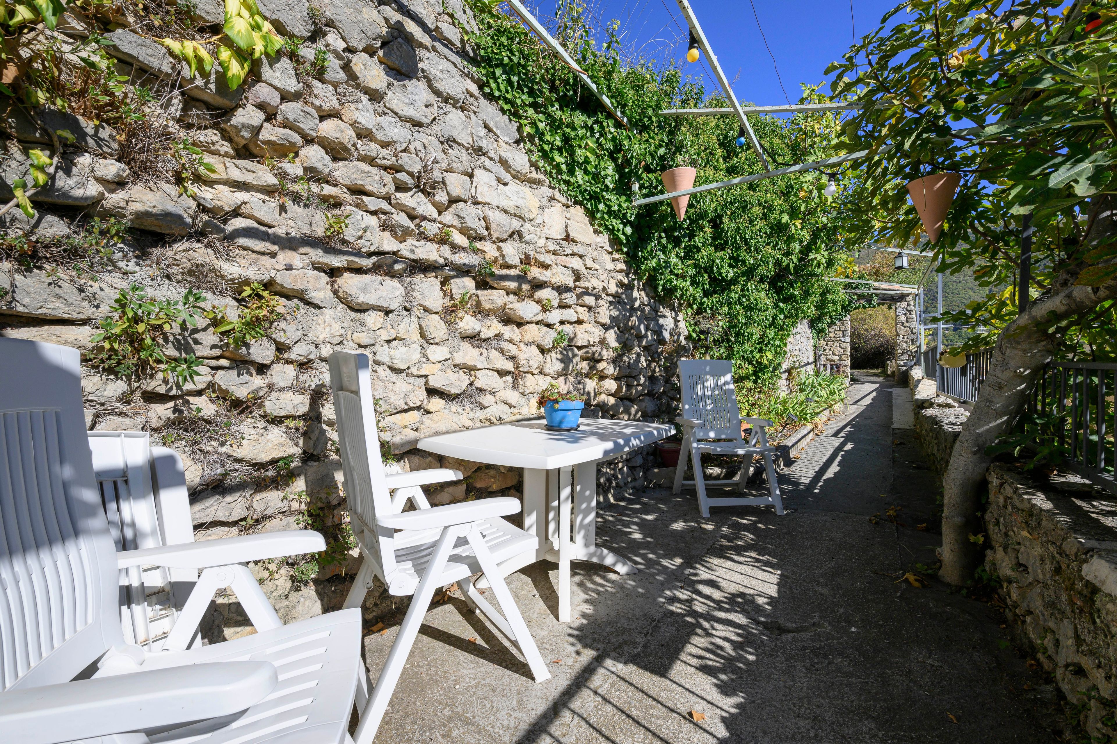 Un mur de pierre bordé d’un jardin agrémenté de chaises et d’une table en plastique blanc, d’un étroit sentier et d’une vue sur une montagne au loin.